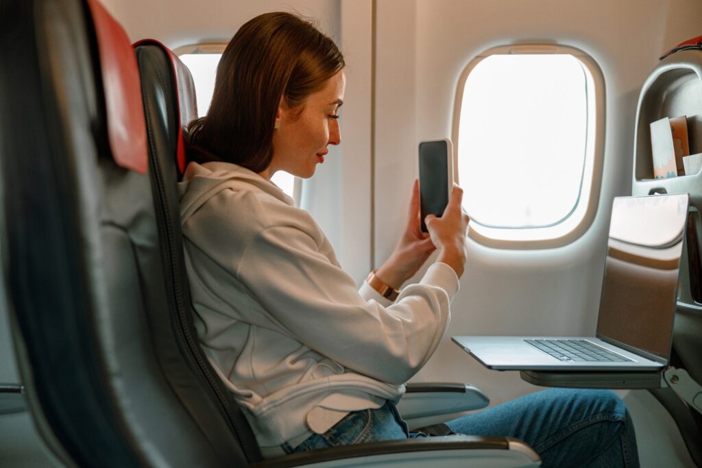 A young woman uses a smartphone and laptop while on a plane.